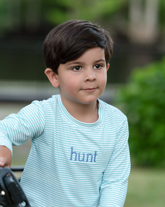 A young boy wearing a light blue and white striped long-sleeve t-shirt with the word 'hunt' embroidered in blue on the chest, photographed outdoors against a soft green background.