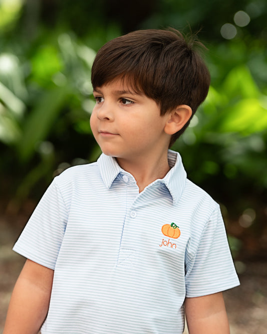 A young boy wearing a light blue and white striped polo shirt with a custom pumpkin and the name 'John' embroidered on the chest, set against a soft, blurred green garden background.