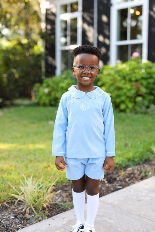 A young boy wearing a light blue long-sleeve collared shirt and matching shorts, standing outdoors on a sidewalk in front of a house and greenery.