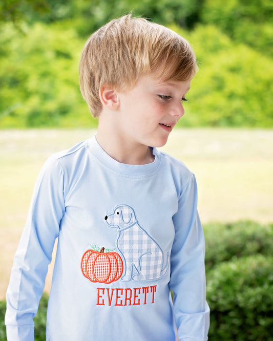 A young boy wearing a light blue long-sleeve shirt with a dog and pumpkin graphic, standing outdoors in front of a blurred green background.