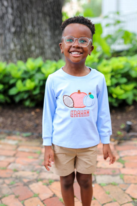 A young boy wearing a light blue long-sleeve t-shirt with a custom fruit graphic and the name 'GRAHAM' printed on the front, paired with khaki shorts, standing outdoors on a brick path with green foliage in the background.