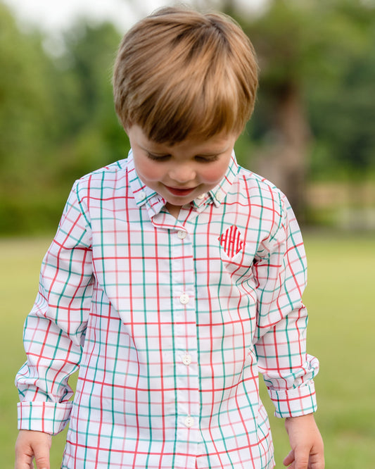 A young boy wearing a long-sleeve button-down shirt with a red, green, and white plaid pattern and a red monogram on the chest, standing outdoors in a grassy park.