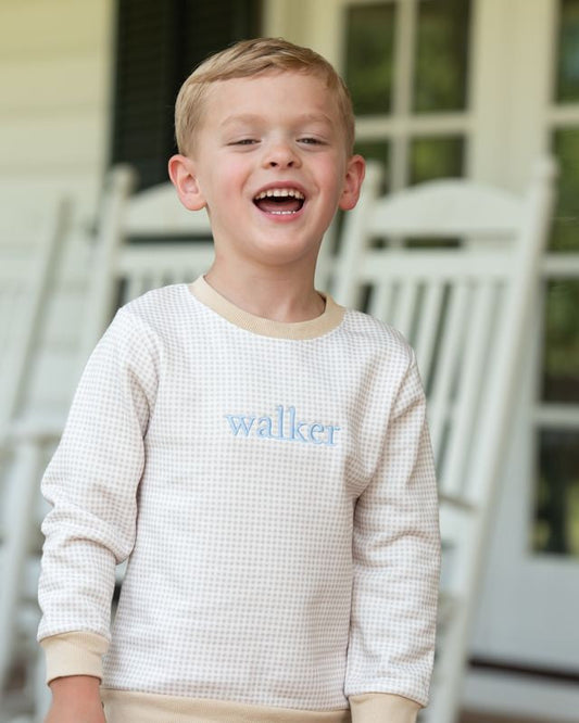 A young boy with blond hair smiling and laughing, wearing a cream waffle-knit long-sleeve shirt with 'walker' embroidered in light blue on the chest, standing on a porch with white rocking chairs in the background.