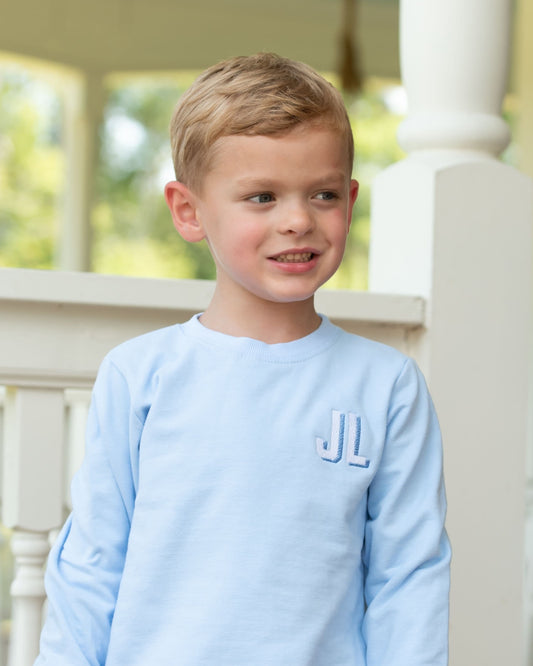 A young boy with blond hair smiling on a porch, wearing a light blue long-sleeve crewneck shirt with a monogrammed 'JL' on the left chest.