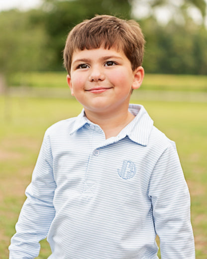 A young boy with brown hair smiling outdoors, wearing a light blue and white striped long-sleeve polo shirt with a monogrammed 'JB' on the chest.