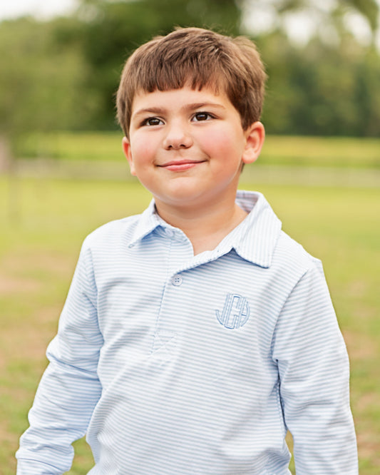A young boy with brown hair smiling outdoors, wearing a light blue and white striped long-sleeve polo shirt with a monogrammed 'JB' on the chest.