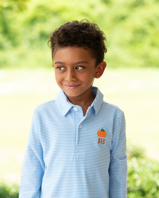 A young boy with curly dark hair wearing a light blue and white striped polo shirt with an orange embroidered logo on the chest, standing outdoors against a soft, blurred green background.