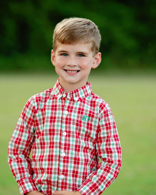 A young boy with light brown hair smiling outdoors, wearing a red, white, and green plaid long-sleeve button-down shirt.
