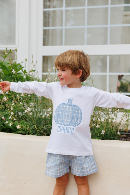 A young boy with light brown hair stands outdoors with his arms outstretched, wearing a white long-sleeve t-shirt with a blue plaid pumpkin graphic, paired with matching plaid shorts.