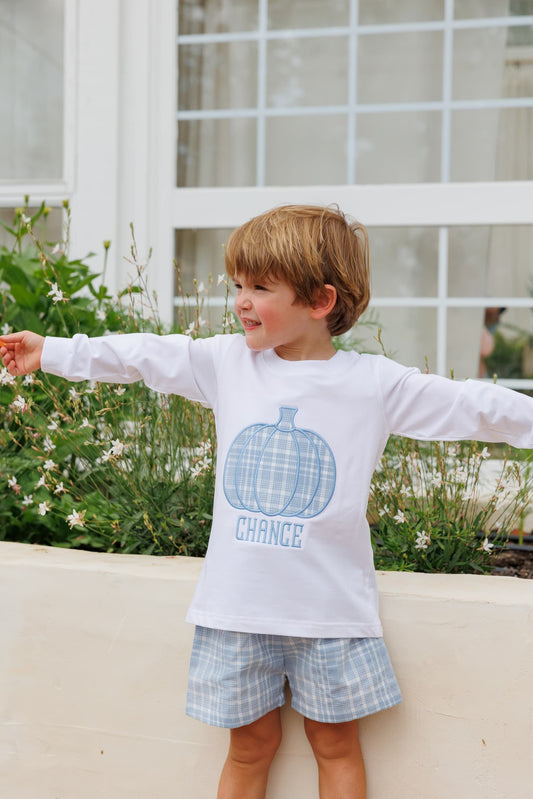 A young boy with light brown hair stands outdoors with his arms outstretched, wearing a white long-sleeve t-shirt with a blue plaid pumpkin graphic, paired with matching plaid shorts.