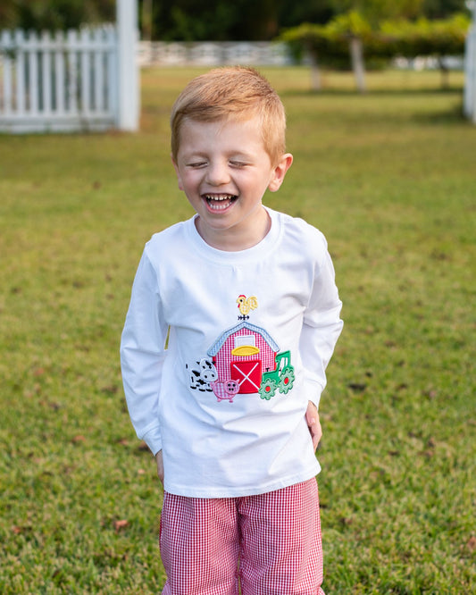 A young boy with light hair is smiling outdoors on a grassy lawn, wearing a white long-sleeve t-shirt with a colorful embroidered barn and tractor graphic on the front, paired with pink and white gingham pants.
