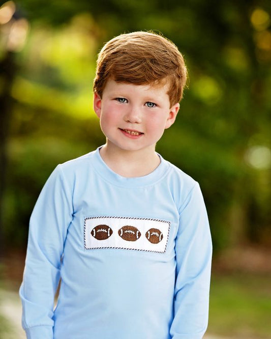 A young boy with red hair wearing a light blue long-sleeve shirt with three brown footballs on a white rectangular patch on the chest, set against a blurred green outdoor background.