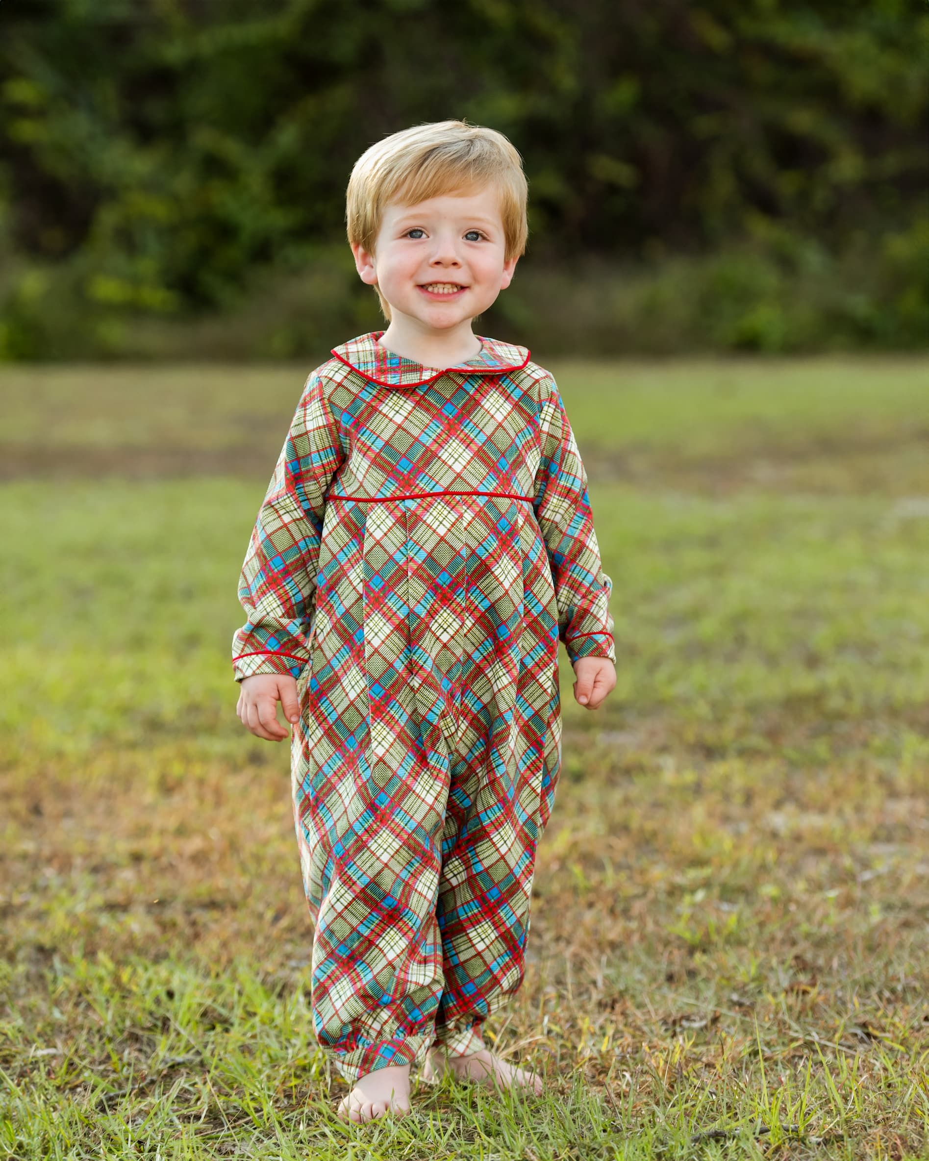 A young child standing barefoot on grass outdoors, wearing a long-sleeve footless plaid jumpsuit in a multicolor tartan pattern with red, blue, green, and beige tones.
