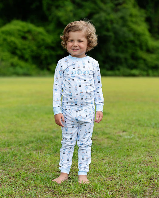 A young child standing outdoors on a grassy lawn wearing a light blue and white long-sleeve pajama set with a repeating architectural pattern.