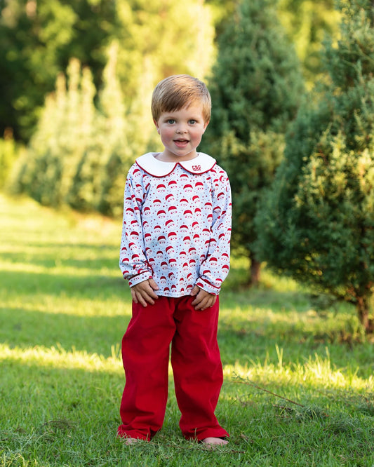A young child standing outdoors on grass in front of evergreen trees, wearing a white long-sleeve top with a red Santa Claus pattern and a white Peter Pan collar, paired with bright red pants.