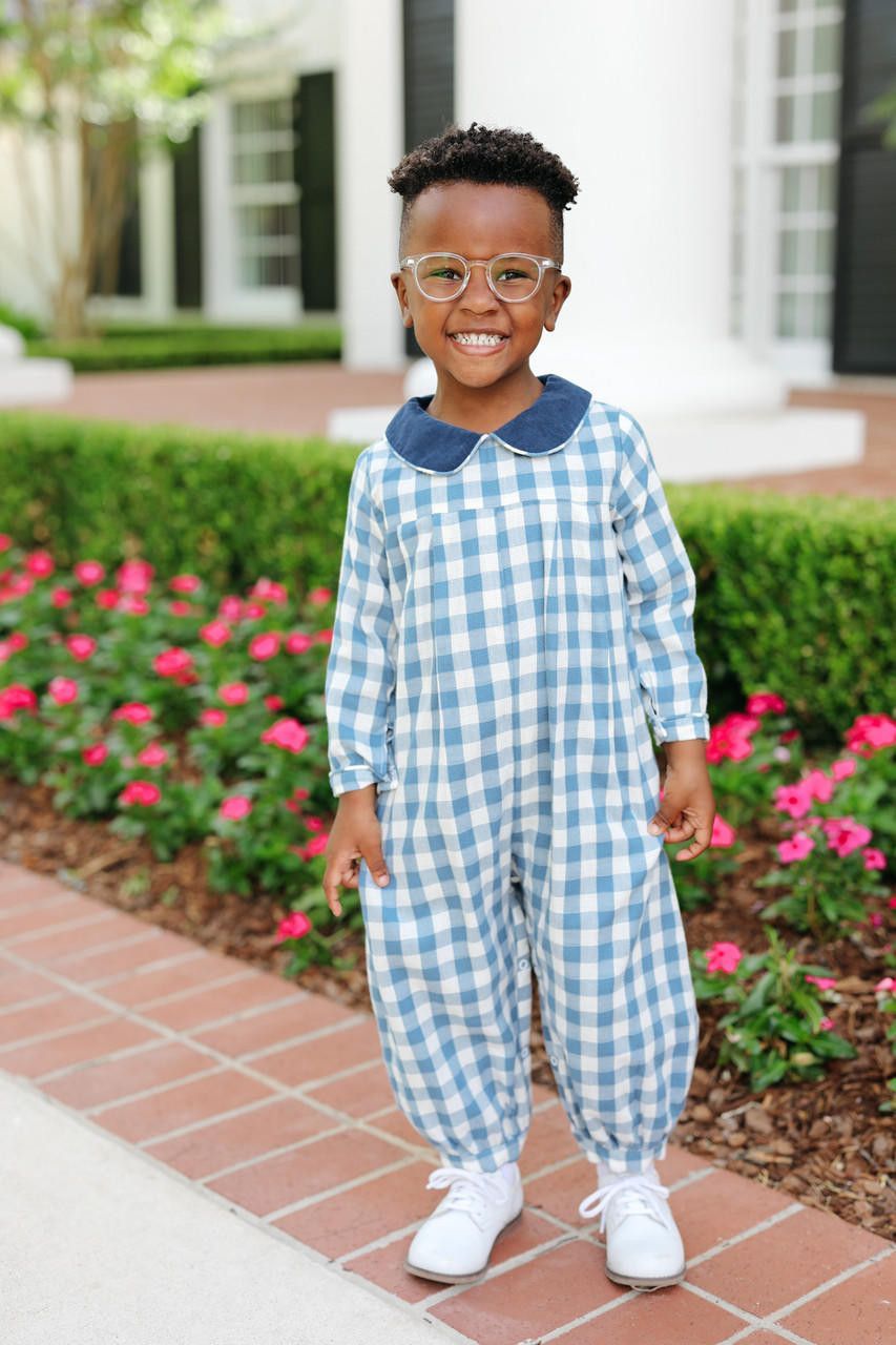 A young child wearing a light blue and white gingham long-sleeve jumpsuit with a dark blue collar, standing outdoors on a brick walkway in front of a house and flower beds.