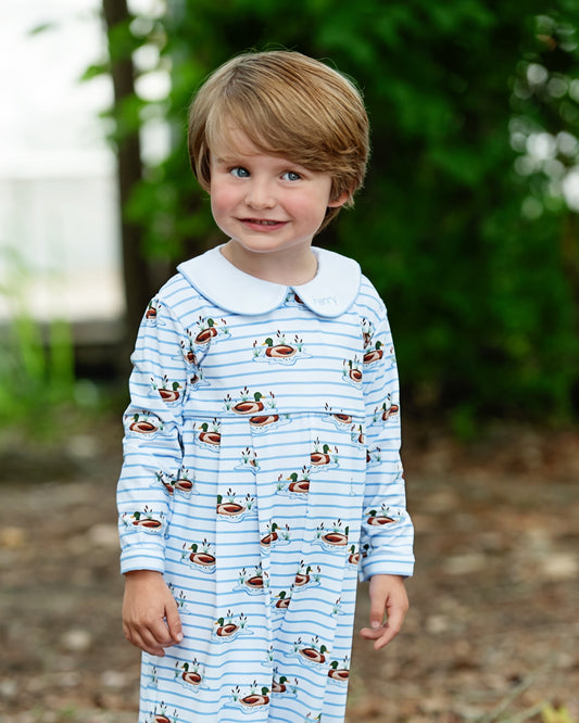 A young child wearing a light blue and white striped long-sleeve nightgown with a white Peter Pan collar and a repeating duck print, standing outdoors against a blurred green background.