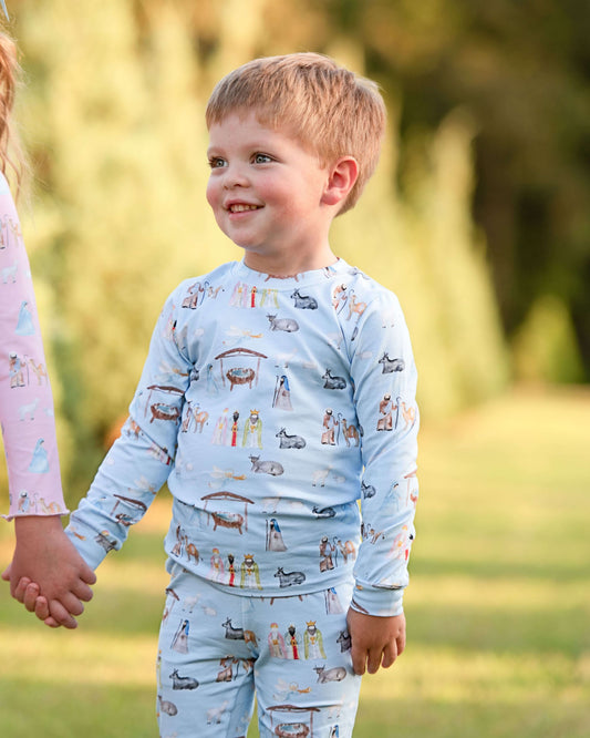 A young child wearing a light blue long-sleeve pajama set with a colorful cartoon animal and house pattern, standing outdoors in a grassy park setting.
