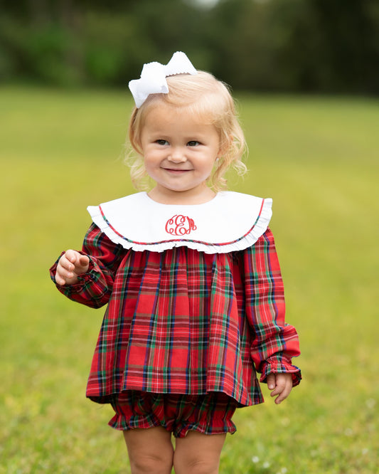 A young child wearing a red and green plaid dress with a white ruffled collar featuring a red monogram, standing outdoors on a grassy field.