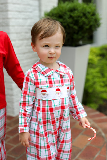 A young child wearing a red, white, and green plaid holiday pajama top with a white waistband featuring embroidered Santa faces, standing outdoors on a brick patio.