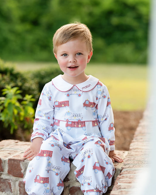 A young child wearing a white long-sleeve pajama set with a red and blue train print, sitting on a brick ledge outdoors.