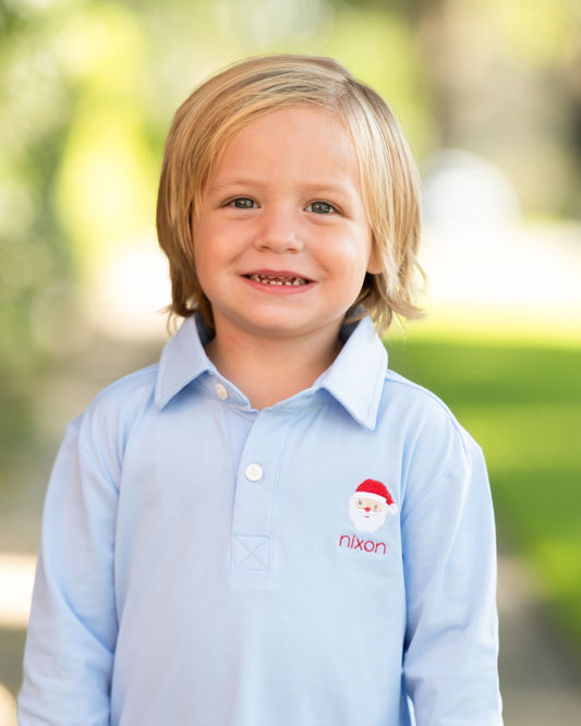 A young child with blonde hair smiling, wearing a light blue long-sleeve polo shirt with a custom embroidered Santa face and the name 'nixon' on the left chest, set against a soft, blurred green outdoor background.