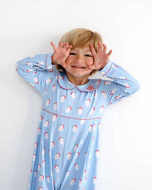 A young child with blonde hair wearing a light blue long-sleeve pajama dress with a white snowman pattern and red trim, posing with their hands around their eyes against a plain white background.