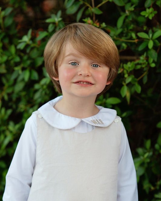 A young child with light brown hair and blue eyes wearing a white long-sleeve shirt with a white Peter Pan collar and a cream-colored textured pinafore dress, standing outdoors in front of a green leafy background.