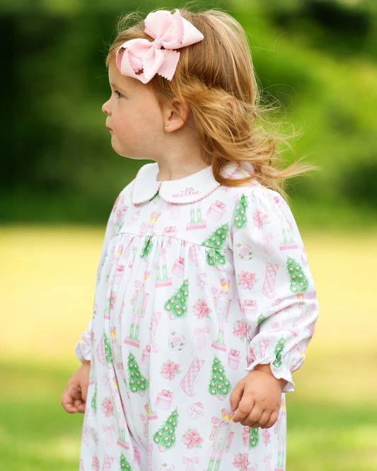 A young girl in profile wearing a white long-sleeve nightgown with a pink and green Christmas print, featuring a Peter Pan collar and a matching pink bow in her hair, set against a soft-focus green outdoor background.