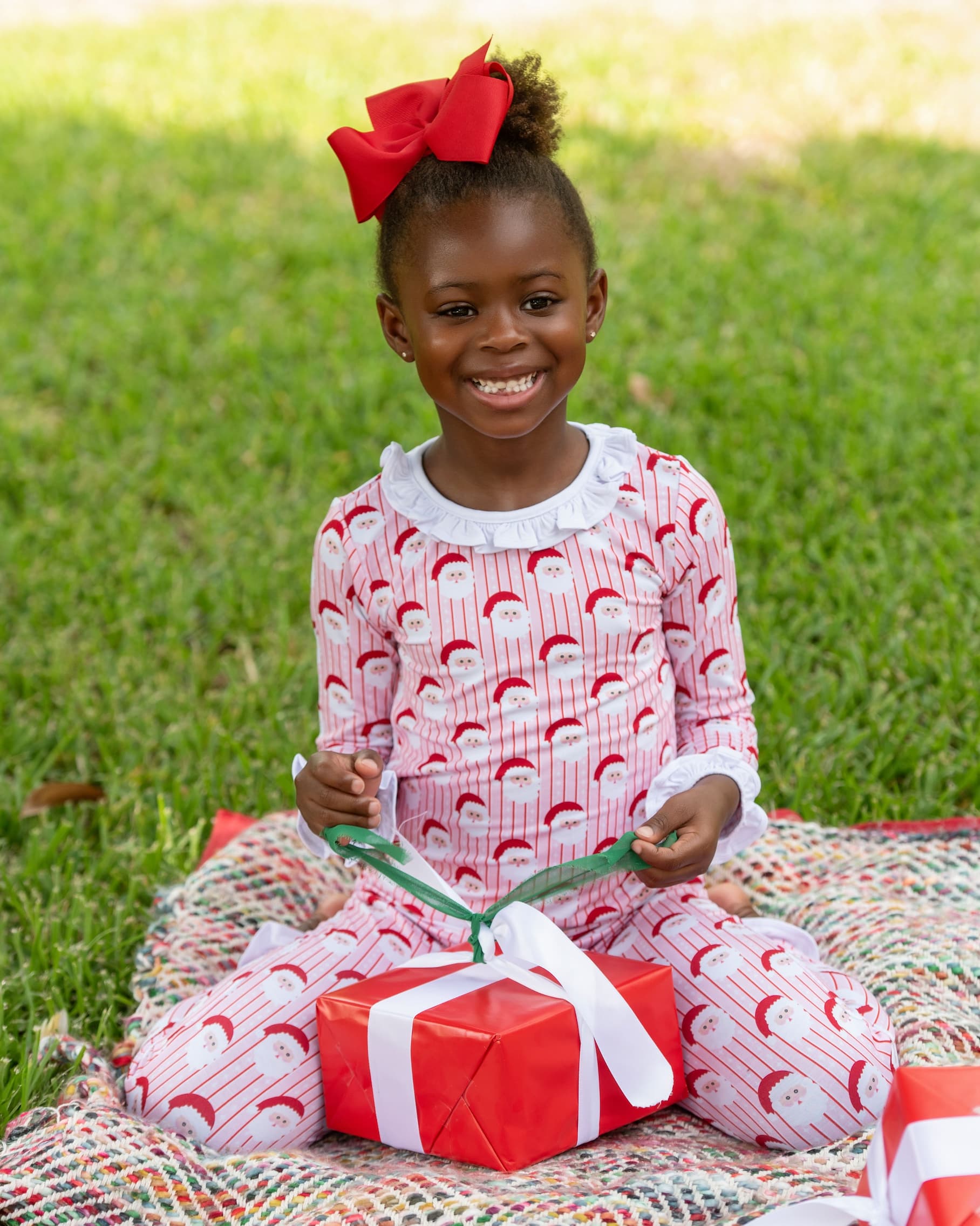 A young girl sitting on a blanket on grass, wearing a red and white holiday-themed pajama set with a matching red bow in her hair. She is smiling and holding a wrapped gift box.