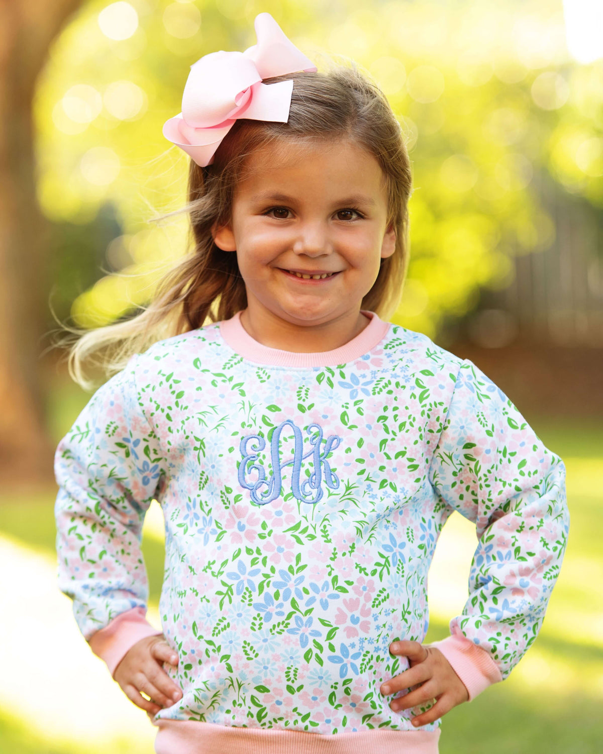 A young girl smiling outdoors wearing a light blue long-sleeve sweatshirt with a pink and green floral pattern, pink trim, and a blue monogram on the chest. She has a large pink bow in her hair, with a blurred green and yellow background.