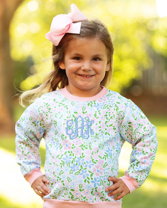 A young girl smiling outdoors wearing a light blue long-sleeve sweatshirt with a pink and green floral pattern, pink trim, and a blue monogram on the chest. She has a large pink bow in her hair, with a blurred green and yellow background.