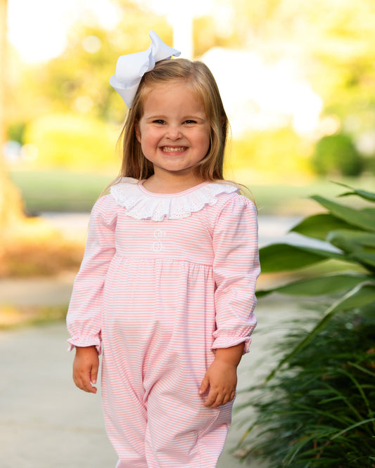 A_young_girl_smiling_outdoors_while_wearing_a_pink_and_white_striped_long-sleeve_romper_with_a_white_ruffled_collar_and_a_white_bow_in_her_hair._1