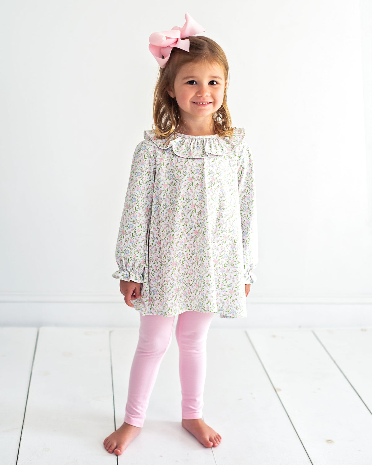 A young girl stands barefoot against a white wall and white floorboards, wearing a long-sleeve white tunic top with a delicate multicolor floral print, light pink leggings, and a large pink bow in her hair.
