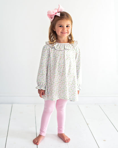 A young girl stands barefoot against a white wall and white floorboards, wearing a long-sleeve white tunic top with a delicate multicolor floral print, light pink leggings, and a large pink bow in her hair.