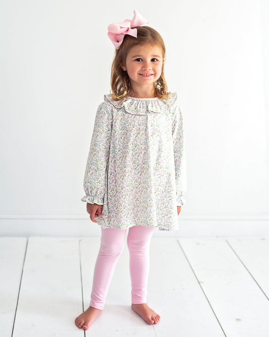 A young girl stands barefoot against a white wall and white floorboards, wearing a long-sleeve white tunic top with a delicate multicolor floral print, light pink leggings, and a large pink bow in her hair.