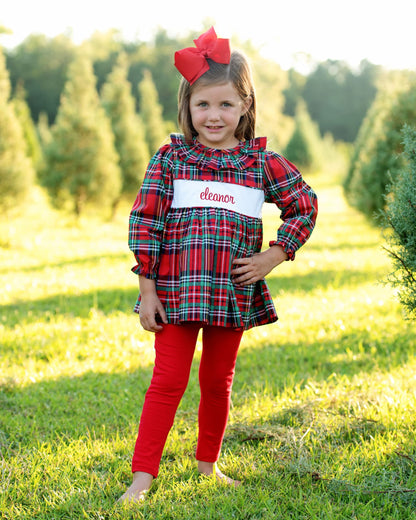 A young girl stands barefoot in a grassy field with trees in the background, wearing a red and green plaid long-sleeve top with a white name patch that reads 'elmer', red leggings, and a large red bow in her hair.