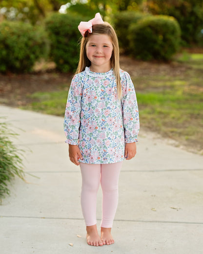 A young girl stands barefoot on a concrete path outdoors, wearing a long-sleeve floral tunic top with a high ruffled collar and light pink leggings. She has a pink bow in her hair.