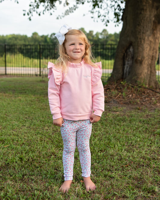 A young girl stands barefoot on a grassy lawn in front of a large tree, wearing a light pink long-sleeve top with ruffle details and white patterned leggings.