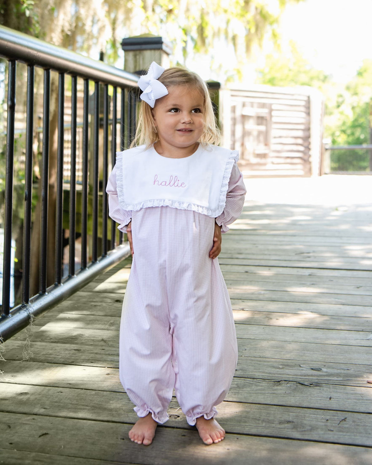 A young girl stands barefoot on a wooden boardwalk wearing a light pink bubble romper with a white bib collar embroidered with the name 'hallee' in pink. She has a white bow in her hair, with trees and a railing in the background.