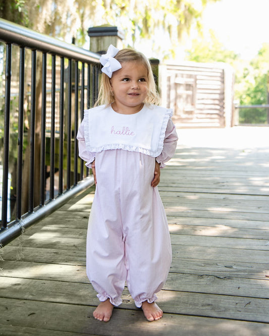 A young girl stands barefoot on a wooden boardwalk wearing a light pink bubble romper with a white bib collar embroidered with the name 'hallee' in pink. She has a white bow in her hair, with trees and a railing in the background.