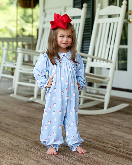 A young girl stands barefoot on a wooden porch wearing a light blue footless one-piece jumpsuit with a penguin print and a large red bow in her hair.
