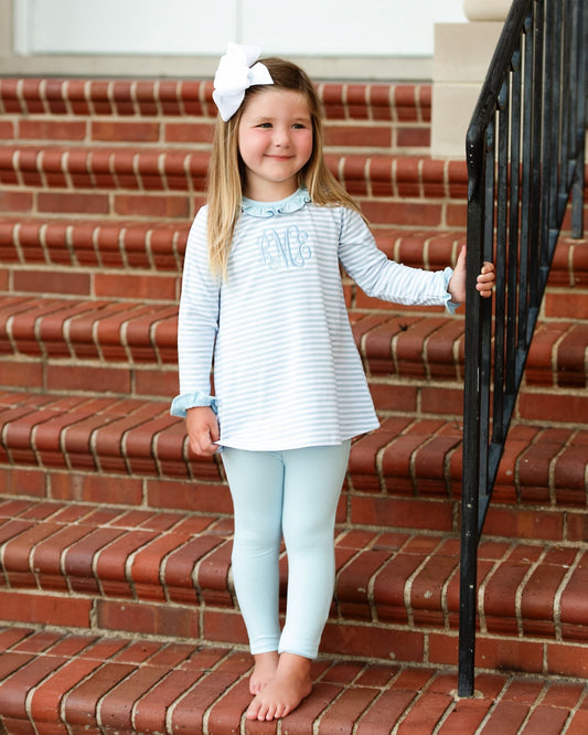 A young girl stands barefoot on red brick stairs, wearing a light blue and white striped long-sleeve top with 'MBS' monogrammed on the chest, light blue leggings, and a white bow in her hair.