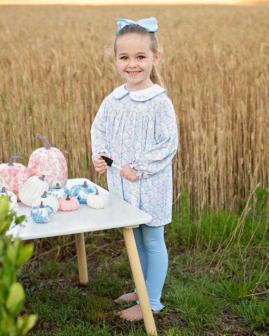A young girl stands barefoot outdoors in a field of wheat next to a small table decorated with pumpkins, wearing a light blue and white patterned long-sleeve dress with a Peter Pan collar and light blue leggings.