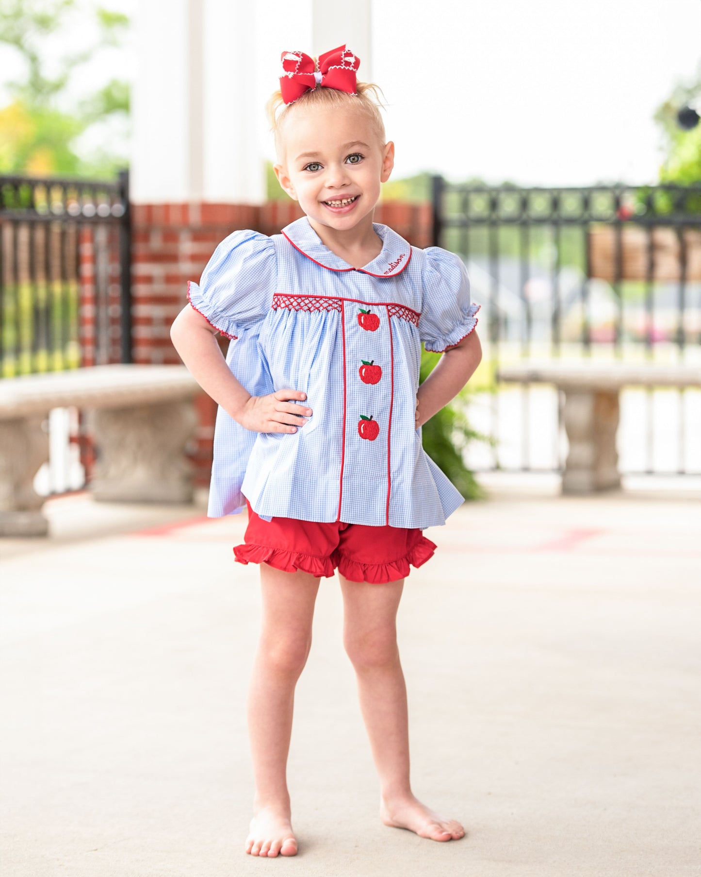 A young girl stands barefoot outdoors wearing a light blue short-sleeve blouse with red trim and apple motifs, red ruffled shorts, and a red bow in her hair.
