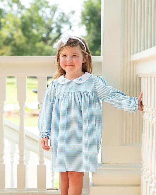 A young girl stands on a white porch wearing a light blue and white striped long-sleeve dress with a white Peter Pan collar decorated with small blue bows. She has a white bow headband and is smiling while looking at the camera.