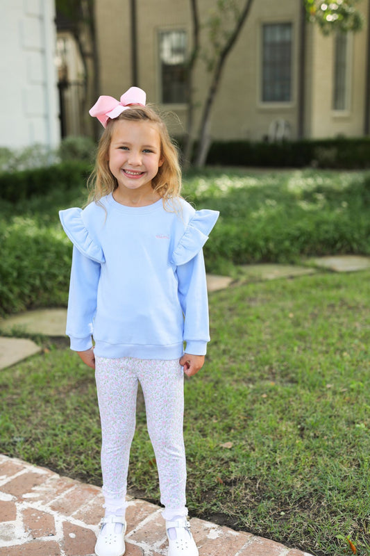 A young girl stands outdoors on a brick path in a grassy yard, wearing a light blue long-sleeve sweatshirt with ruffled shoulder details, white patterned leggings, and white sneakers. She has a pink bow in her hair.