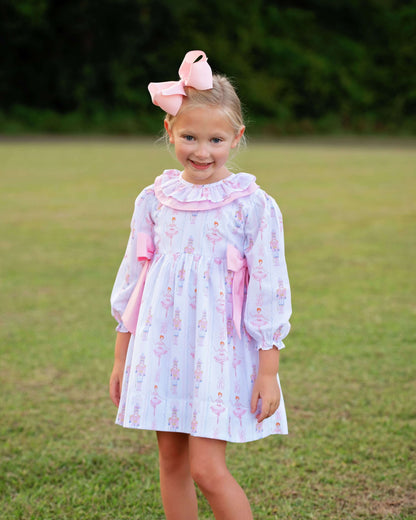 A young girl stands outdoors on a grassy lawn wearing a light lavender and pink ballerina-print dress with a ruffled collar, long sleeves, and matching pink bows, along with a large pink bow in her hair.