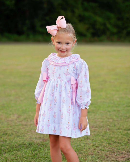 A young girl stands outdoors on a grassy lawn wearing a light lavender and pink ballerina-print dress with a ruffled collar, long sleeves, and matching pink bows, along with a large pink bow in her hair.