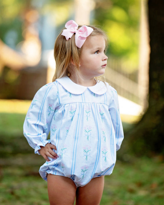 A young girl wearing a light blue and white striped long-sleeve romper with a white Peter Pan collar, standing outdoors in a park-like setting with a pink bow in her hair.